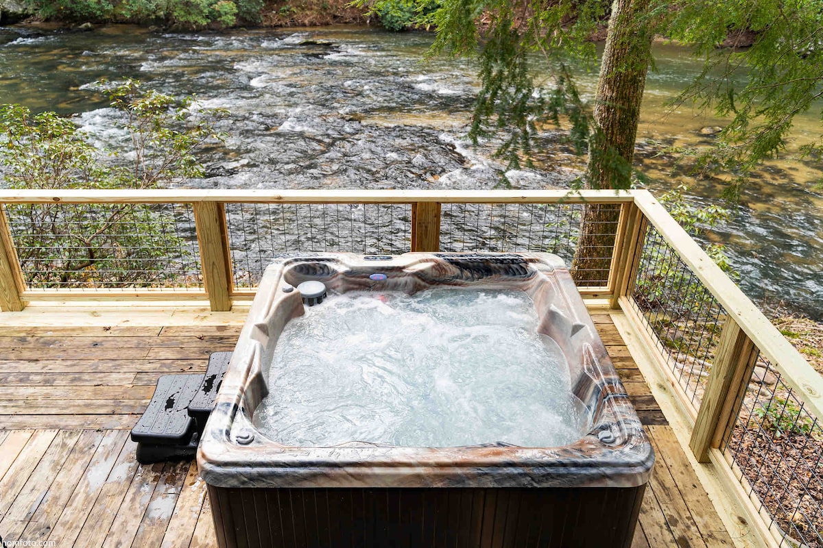 A hot tub is situated on a wooden deck overlooking the flowing Cartecay River. The area is surrounded by lush greenery, enhancing the natural feel of the space. Water bubbles gently within the hot tub, inviting a sense of relaxation and tranquility.