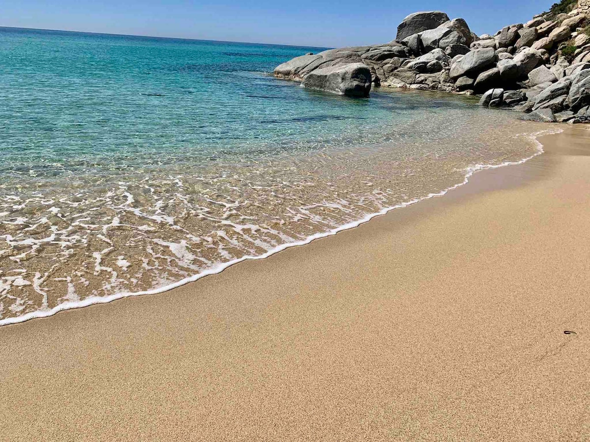 The image captures a serene beach scene with soft golden sand gently meeting a clear turquoise sea. Rounded rocks can be seen in the background, contrasting with the smooth shoreline. The quiet waves create a subtle frothy edge where the water meets the sand.