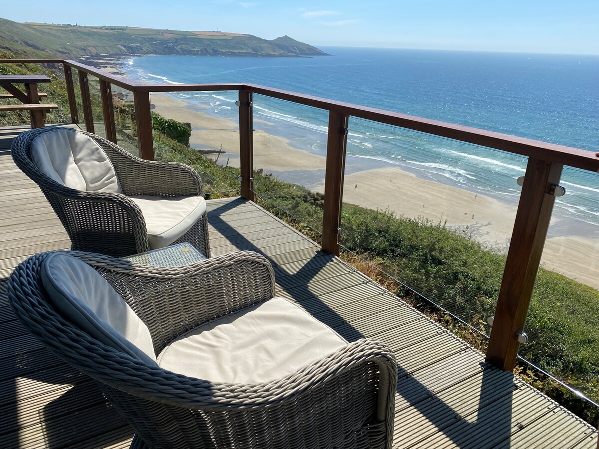Two wicker chairs with soft cushions are positioned on a deck overlooking a sandy beach and the ocean. The railing offers unobstructed views of the coastline and waves, with greenery surrounding the area. Bright sunlight illuminates the scene on a clear day.
