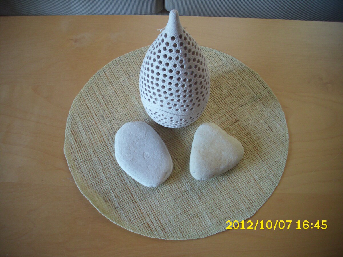 A table is topped with a round, light-colored mat featuring a textured design. Centered is a decorative ceramic object with a pierced pattern, flanked by two smooth, white stones. The composition emphasizes natural elements and soothing colors.