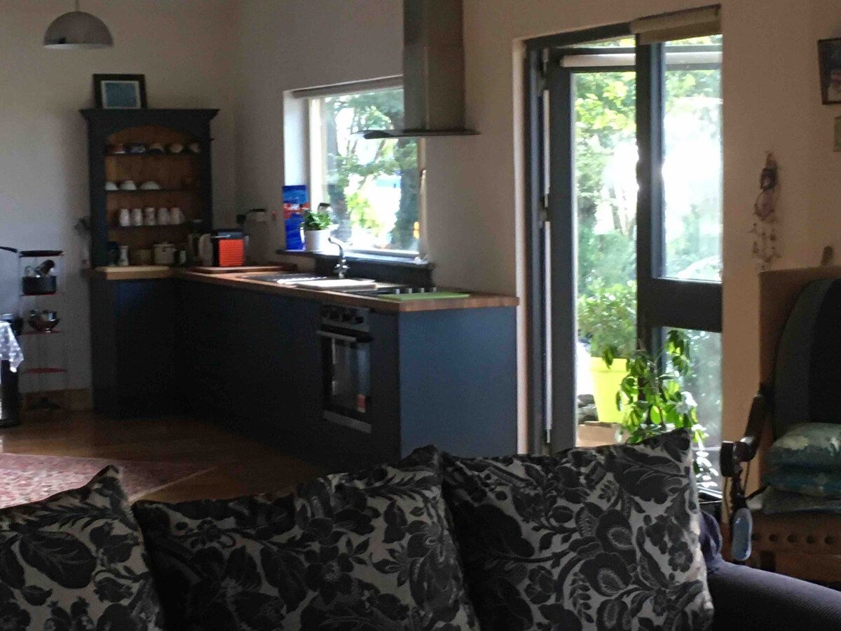 A spacious kitchen area features a well-appointed countertop and modern appliances. Natural light filters in through large windows, illuminating the warm-toned decor and creating a welcoming environment. A comfortable seating area with a floral-patterned couch invites relaxation beside the open doorway leading to the outdoors.