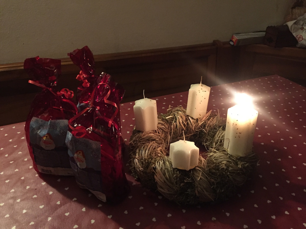 Two red-wrapped gifts are presented alongside a candle arrangement. A circular wreath of natural materials holds five white candles, some of which are lit, casting a soft glow. The background features a table with a light-colored tablecloth adorned with small white patterns.