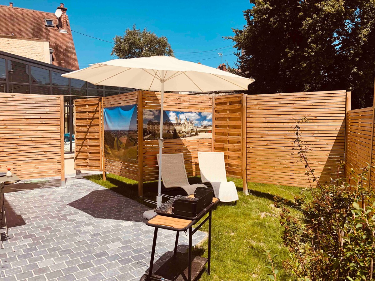 A private patio area is enclosed by wooden slats, providing a sense of seclusion. Two lounge chairs are positioned beneath a large umbrella, with a small table nearby. A green lawn and paved stones create a welcoming outdoor space, perfect for relaxation.