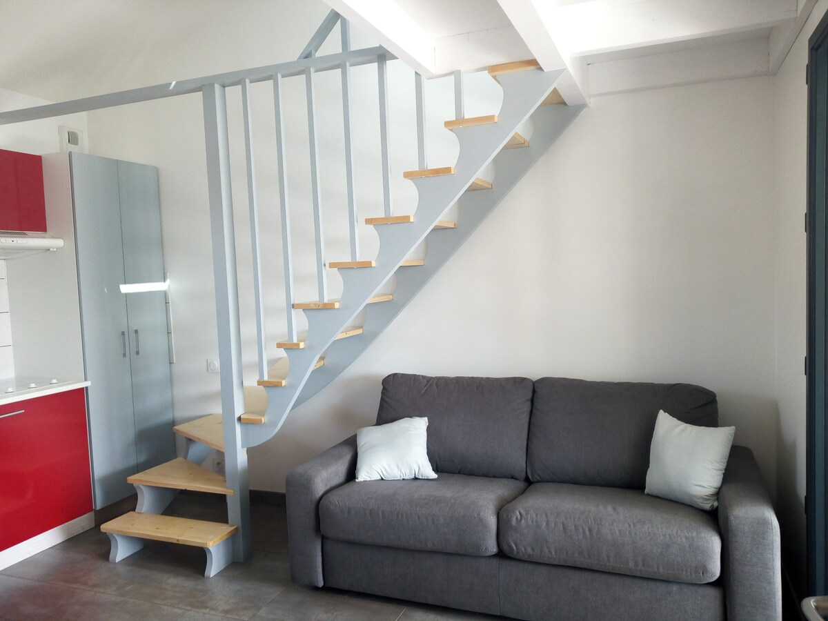 A compact living area features a convertible two-seat sofa with two light-colored cushions. A modern kitchenette with red cabinetry and a white countertop is visible in the background. A light wood staircase leads to a mezzanine area, enhancing the studio's vertical space.