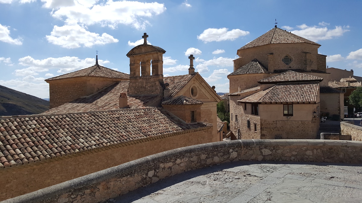 An aerial view captures historic buildings with stone walls and tiled roofs under a bright sky. The architecture includes bell towers and domed roofs, creating a blend of textures and forms. Surrounding landscapes of hills add to the serene ambiance.