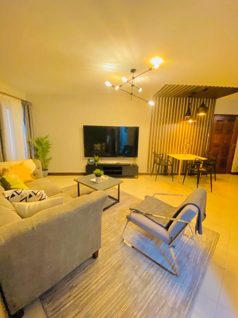 A bright living area features a comfortable gray sofa and a sleek coffee table, set on a textured rug. A large television is mounted on the wall, while a dining nook with a small table and chairs can be seen in the background. Decorative lighting enhances the ambiance.