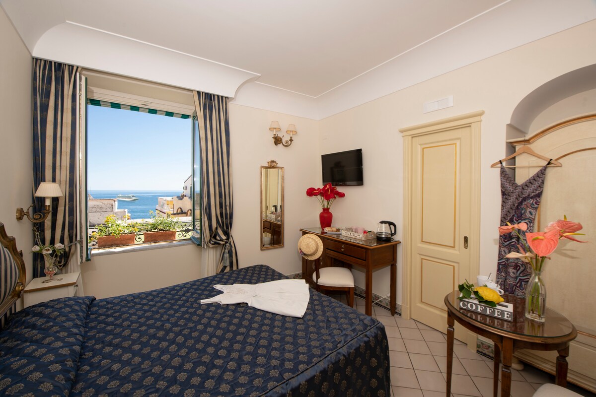 A bright bedroom showcases a spacious bed adorned with navy and gold bedding. A window with a partial sea view allows natural light to fill the room. A wooden desk and a coffee station are positioned alongside a vintage mirror, adding charm to the decor.