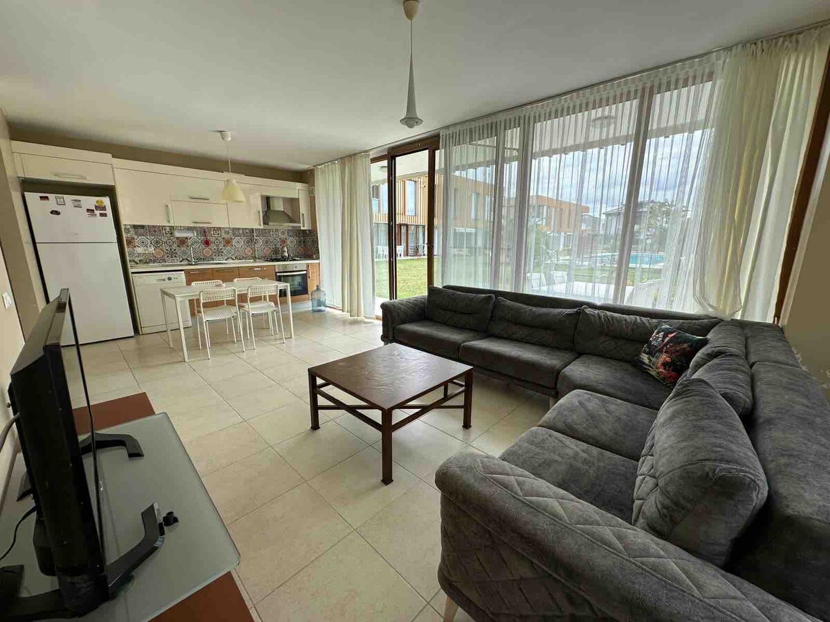 A spacious living area features large windows allowing natural light to fill the room. A comfortable sectional sofa and a wooden coffee table are positioned centrally. The adjoining kitchen area includes white cabinetry and a dining table with chairs, offering a cohesive and open layout.
