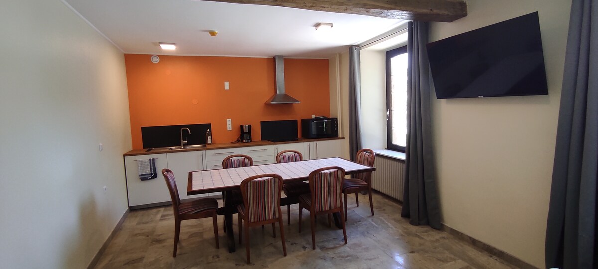 A bright kitchen area is visible, featuring a long dining table with eight chairs. The walls are painted in a warm orange tone, contrasted by dark countertops and modern appliances, including a stovetop and microwave. Natural light enters through a large window, enhancing the inviting space.