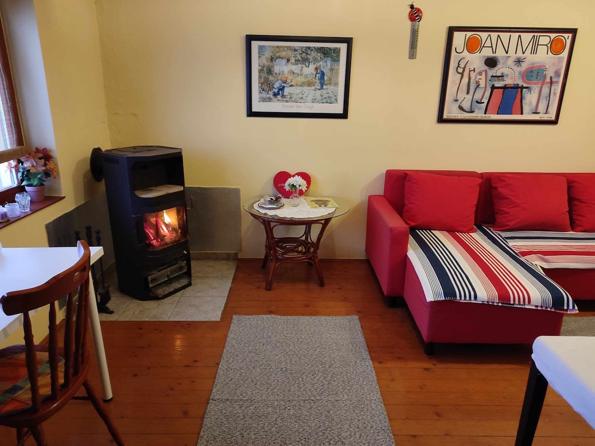 A cozy living space features a red sectional sofa and a small round table with decorative items. A wood-burning stove is positioned against the wall, providing warmth. Art pieces adorn the walls, while natural light enters through a window with wooden frames.