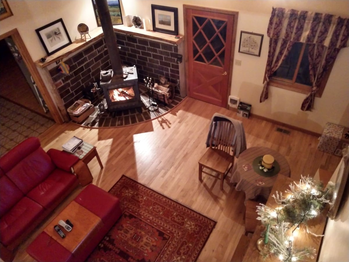A spacious living area features a wood stove with a fire, accompanied by a red sofa and a small dining table. The warm wooden floors are complemented by an area rug. Artworks decorate the walls, and a window with patterned curtains allows light to enter.