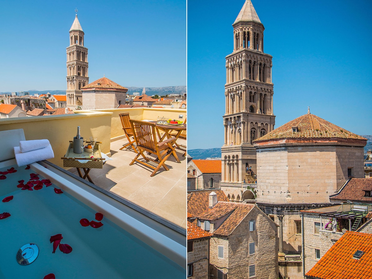 A private terrace is presented, featuring a sunlit area with wooden chairs and a table, set against a backdrop of the Saint Domnius bell tower. A bathtub filled with water and adorned with rose petals adds a romantic touch, enhancing the scenic view of the historic architecture.