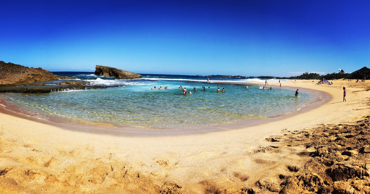A sandy beach stretches along a calm cove, where gentle waves lap the shore. Several individuals can be seen enjoying the water, with rocky formations visible in the distance. The clear blue sky enhances the serene atmosphere, creating an inviting setting for relaxation.