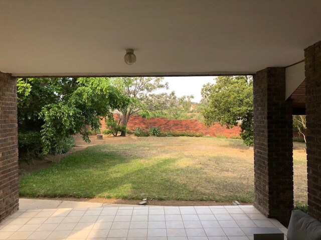 A covered outdoor space leads to a spacious garden area, framed by greenery and trees. The well-maintained lawn is viewed through wide openings, providing a sense of tranquility. Natural light filters through, illuminating the tiled floor and surrounding landscape.