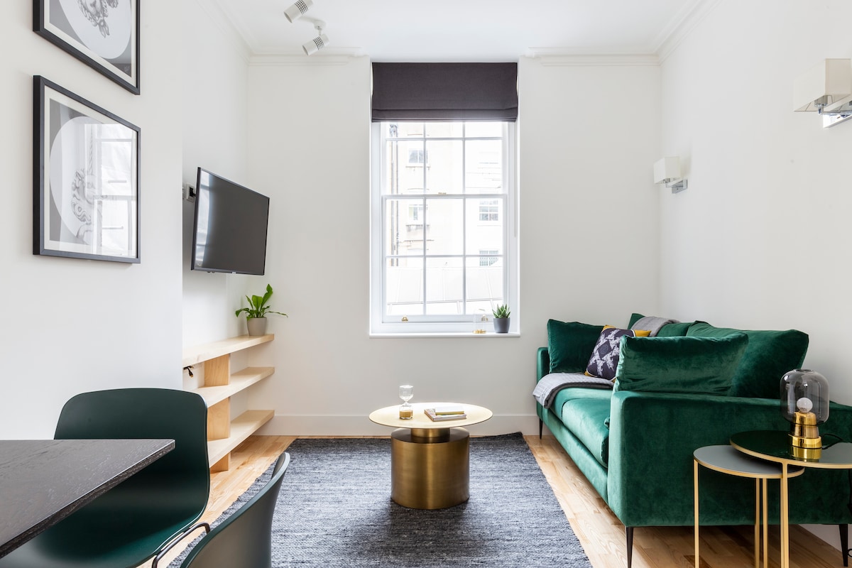 An open plan living area features a comfortable green sofa paired with a stylish round coffee table. A wall-mounted Smart TV is positioned for easy viewing, and a light rug covers the wooden floor. Natural light enters through a large window with a dark shade.