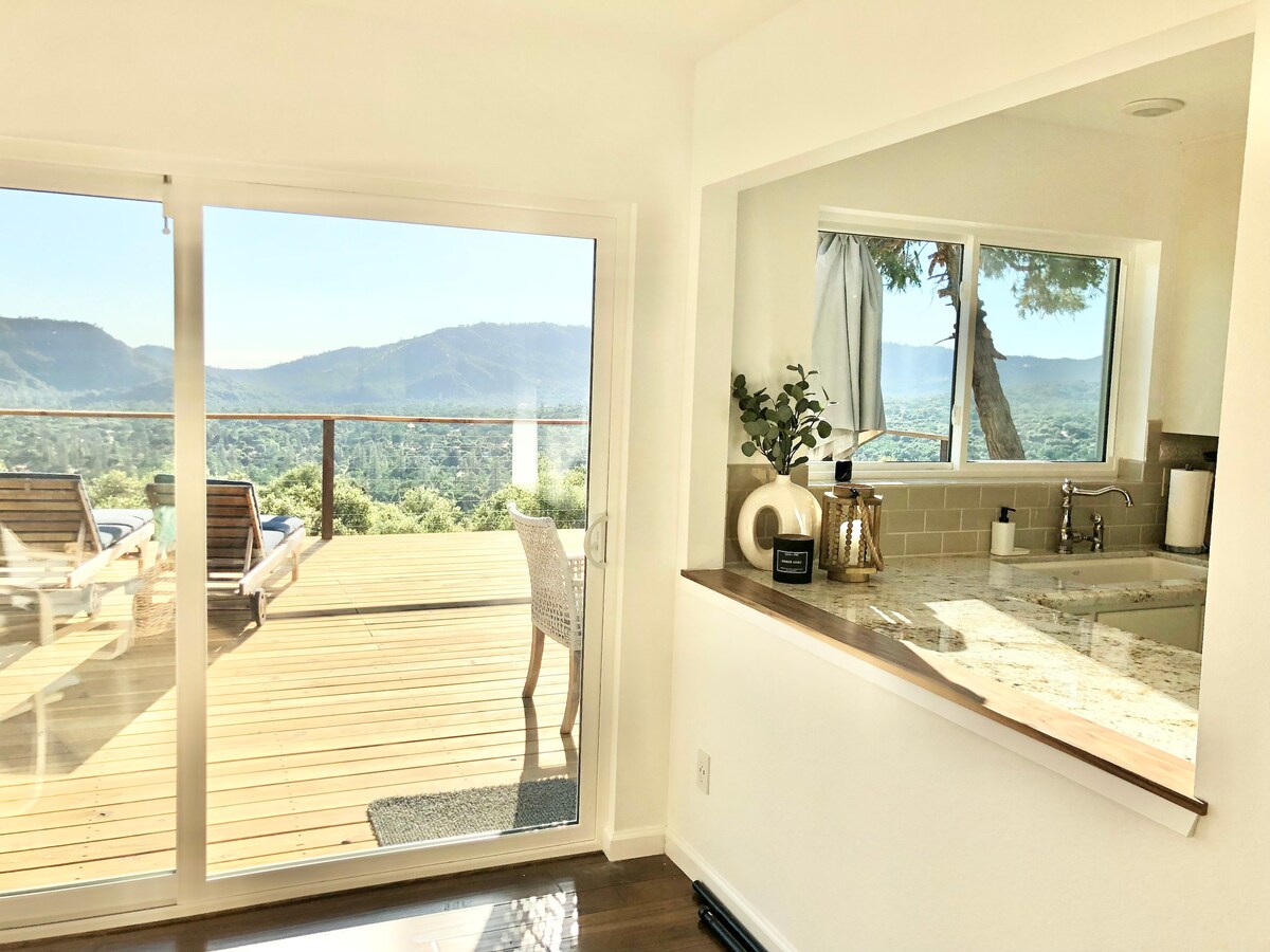 A bright kitchen area with a countertop features modern appliances and overlooks a spacious outdoor deck. Large glass doors provide a clear view of panoramic mountain landscapes, inviting natural light into the room. Outdoor seating is visible on the deck, enhancing the connection with nature.