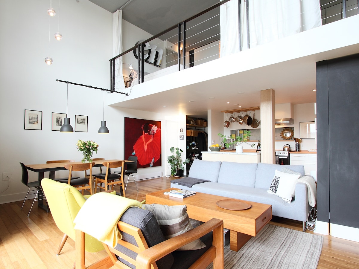 The loft's open living area features mid-century modern furniture, including a light grey sofa and a wooden coffee table. A dining table with chairs is visible, along with framed art on the walls. The kitchen is integrated into the space, showcasing contemporary design elements.