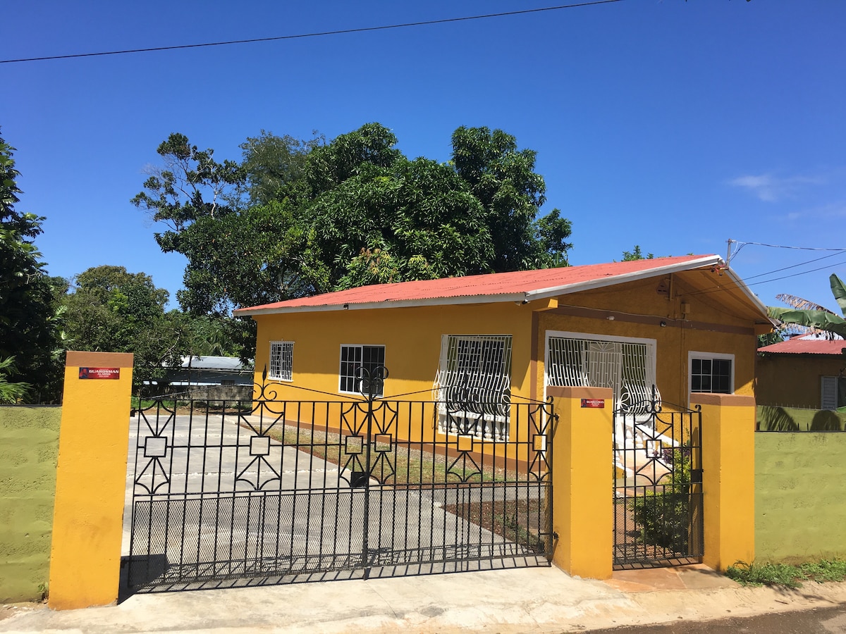 A bright yellow vacation home stands behind a secure, gated entrance. Surrounding greenery provides a natural backdrop, while a paved driveway leads to the front of the house. Large windows allow for ample light, and the roof features a subtle overhang protecting the entrance.