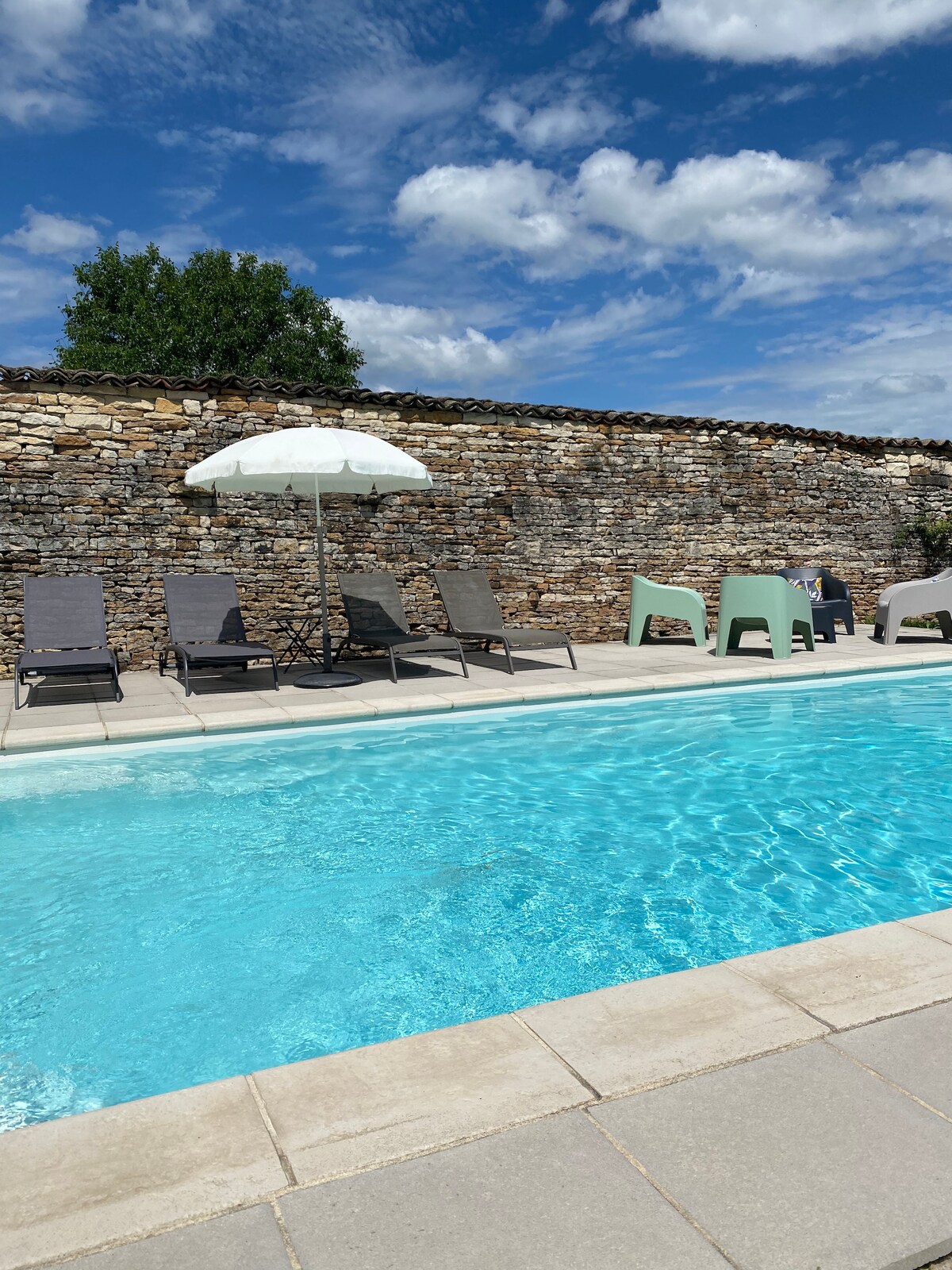 The outdoor pool area features a clear blue pool surrounded by stone walls. Multiple sun loungers are positioned for relaxation, with a large umbrella providing shade. A couple of tables are arranged nearby, complementing the inviting space for outdoor enjoyment.
