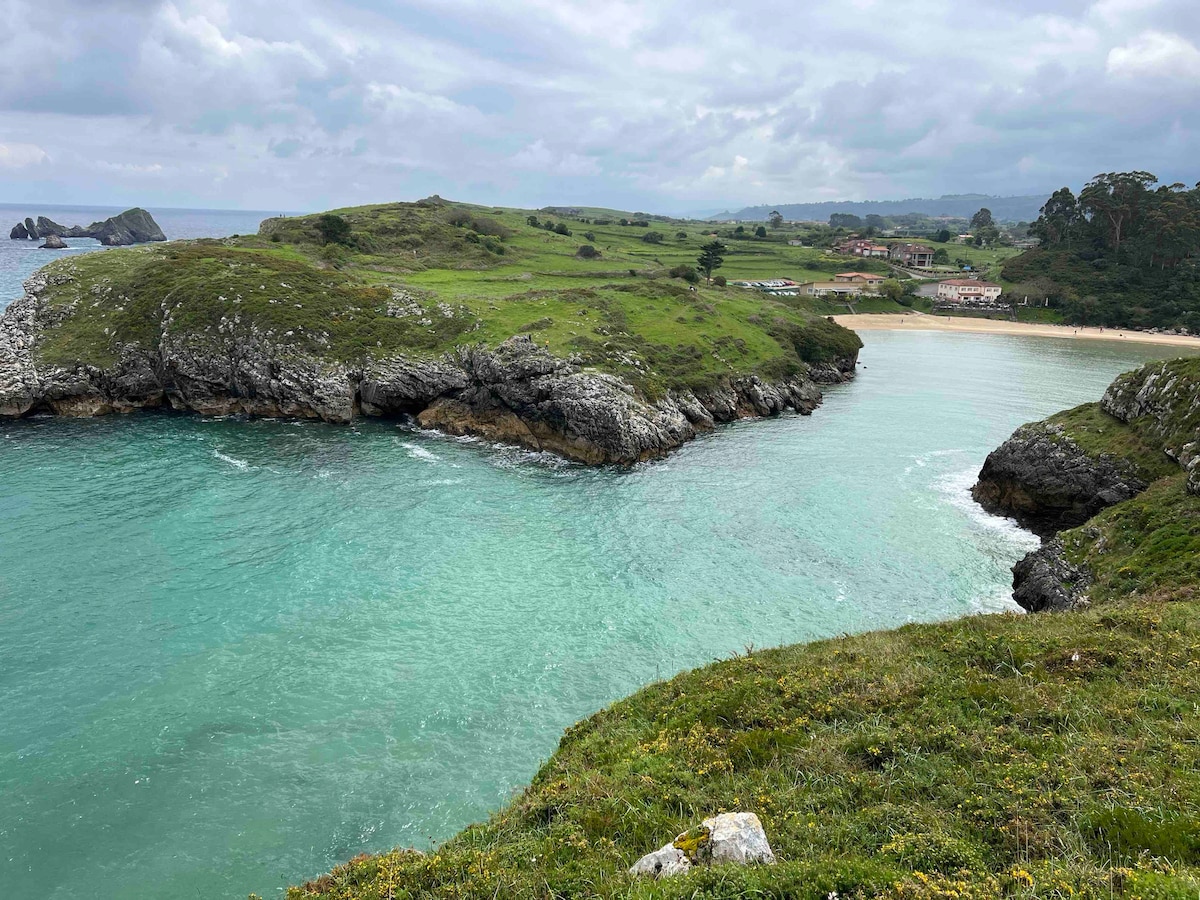 A serene coastal landscape features turquoise waters gently flowing between rocky outcrops. Lush green hills rise in the background, leading to a sandy beach. The scene is framed by a partially cloudy sky, creating a peaceful atmosphere ideal for relaxation.