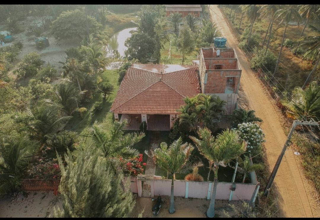 An aerial view showcases a red-tiled roof house surrounded by lush greenery and palm trees. A sandy path runs beside the property, which is bordered by a low wall featuring colorful flowers. A serene pond is visible in the background.