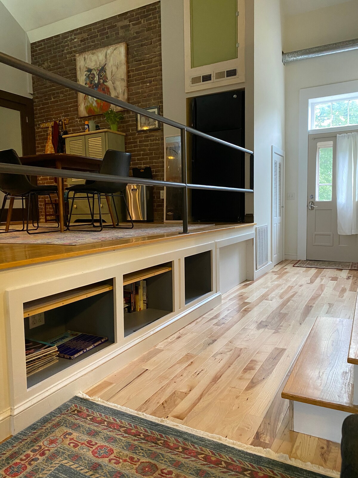 A spacious area is depicted featuring warm wooden flooring and a low, built-in shelf with books visible. A dining table with black chairs is positioned against a textured brick wall. Natural light enters through a large window, complementing the open layout.