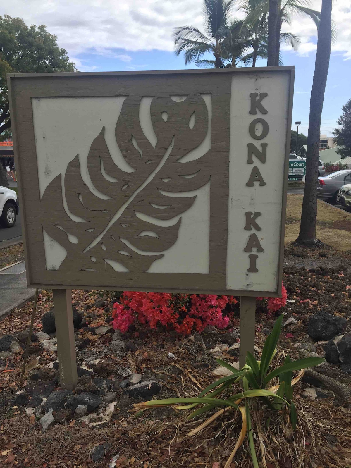 A decorative sign featuring a cut-out leaf design is displayed prominently, indicating the entrance to Kona Kai. The sign is framed in wood and sits among lush landscaping, with flowers blooming in shades of pink and green surrounding its base.