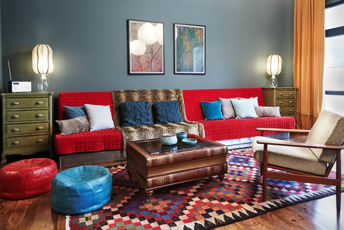 A spacious living room features a vibrant red sofa adorned with various cushions, complemented by a wooden coffee table at the center. Two stylish lamps provide soft lighting, and colorful poufs are arranged on a patterned area rug. Artwork hangs on the blue wall, enhancing the inviting atmosphere.