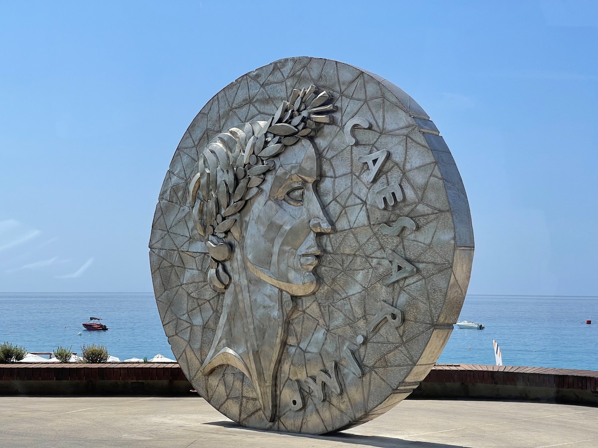 A large, circular sculpture resembling an ancient coin is prominently displayed along the waterfront. The sculpture features intricate geometric patterns and a central figure, framed against a backdrop of clear blue skies and serene ocean waters.
