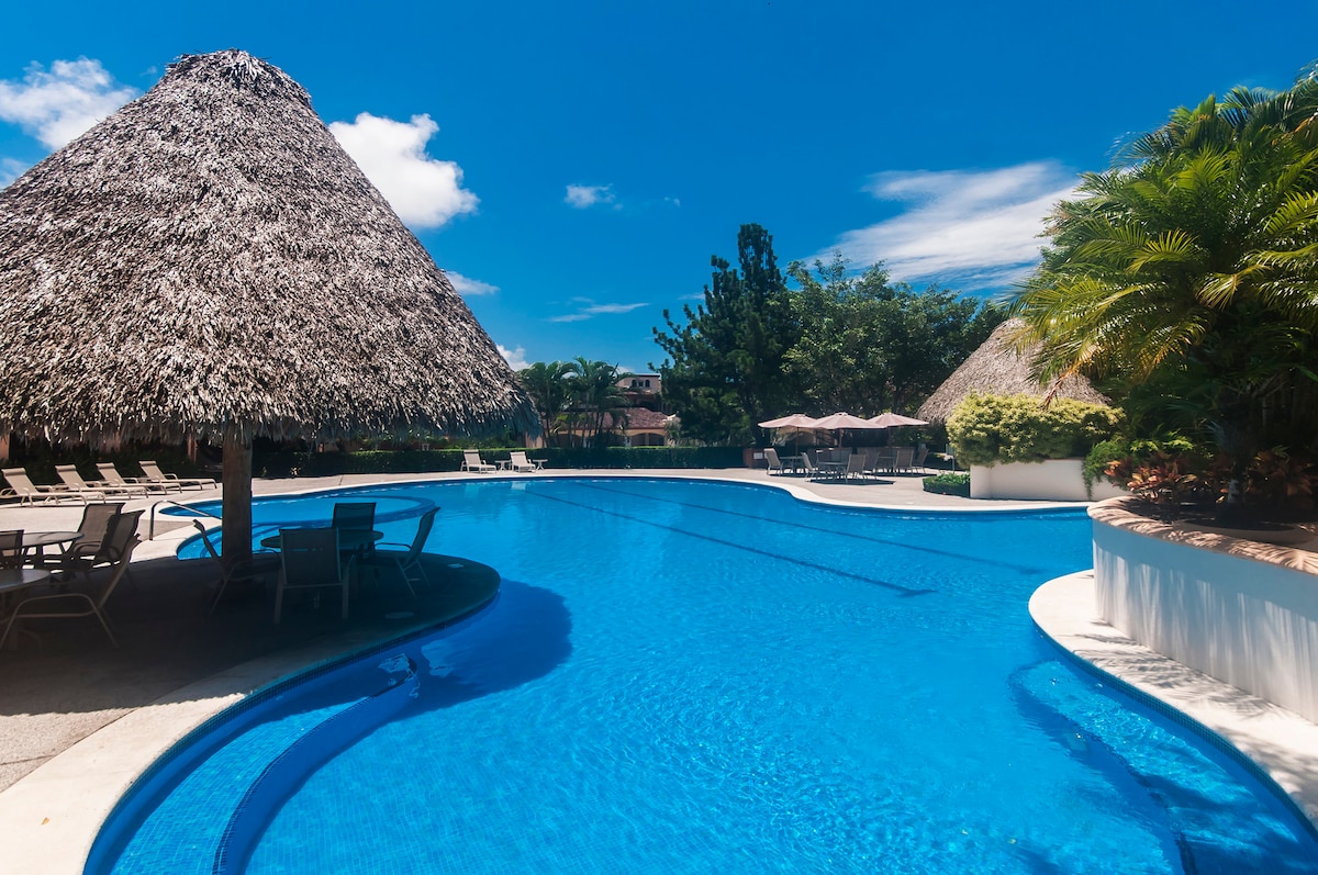A large swimming pool features a clear blue surface, surrounded by lush greenery and tropical landscaping. Thatched-roof cabanas provide shaded seating areas, while sun loungers line the poolside. The bright sky enhances the inviting ambiance of this outdoor relaxation space.