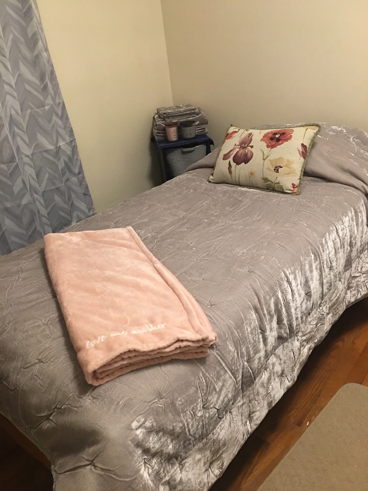 A cozy sleeping space is presented with a single bed covered in a soft gray comforter. A decorative pillow adds a floral accent, while a light pink throw blanket is neatly folded at the foot of the bed. Soft curtains partially frame the window, contributing to a warm ambiance.