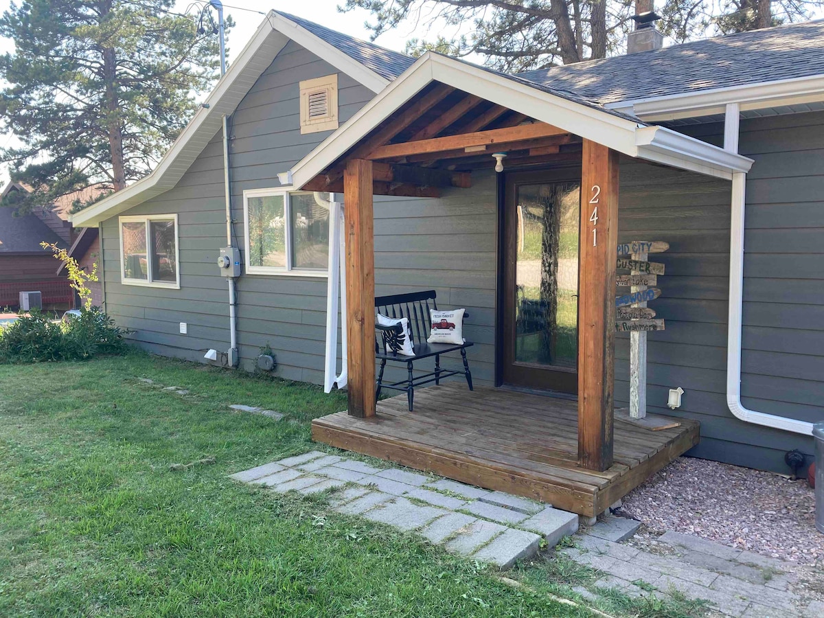 A welcoming porch area features a wooden entryway with a black bench adorned with a decorative pillow. The exterior of the home is clad in gray siding, with multiple windows allowing natural light. Nearby, a stone path leads through a grassy yard.