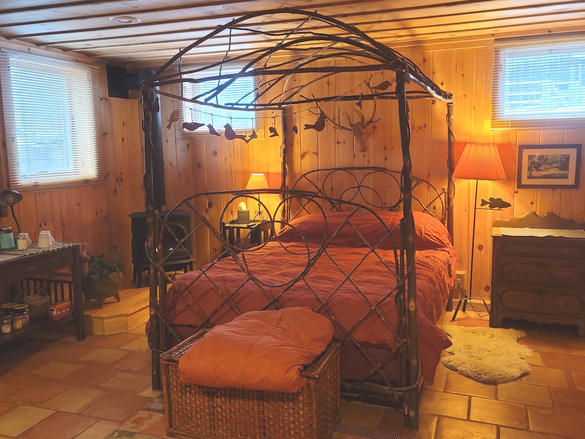 A welcoming bedroom features a decorative wrought iron canopy bed with warm orange bedding. Soft lighting from two lamps creates a cozy atmosphere, while wooden walls and a tiled floor add rustic charm. A wicker basket is placed at the foot of the bed, enhancing the room’s functionality.