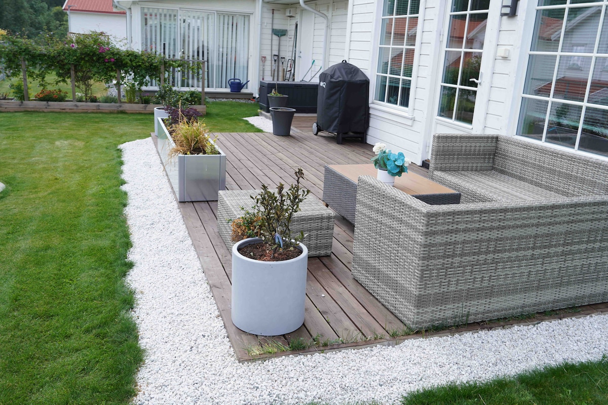 An outdoor veranda is presented with a wooden deck featuring a seating arrangement made of textured, light-colored wicker. Potted plants and a small table are arranged alongside planters filled with greenery. A barbecue grill is positioned at the back, providing additional cooking options.
