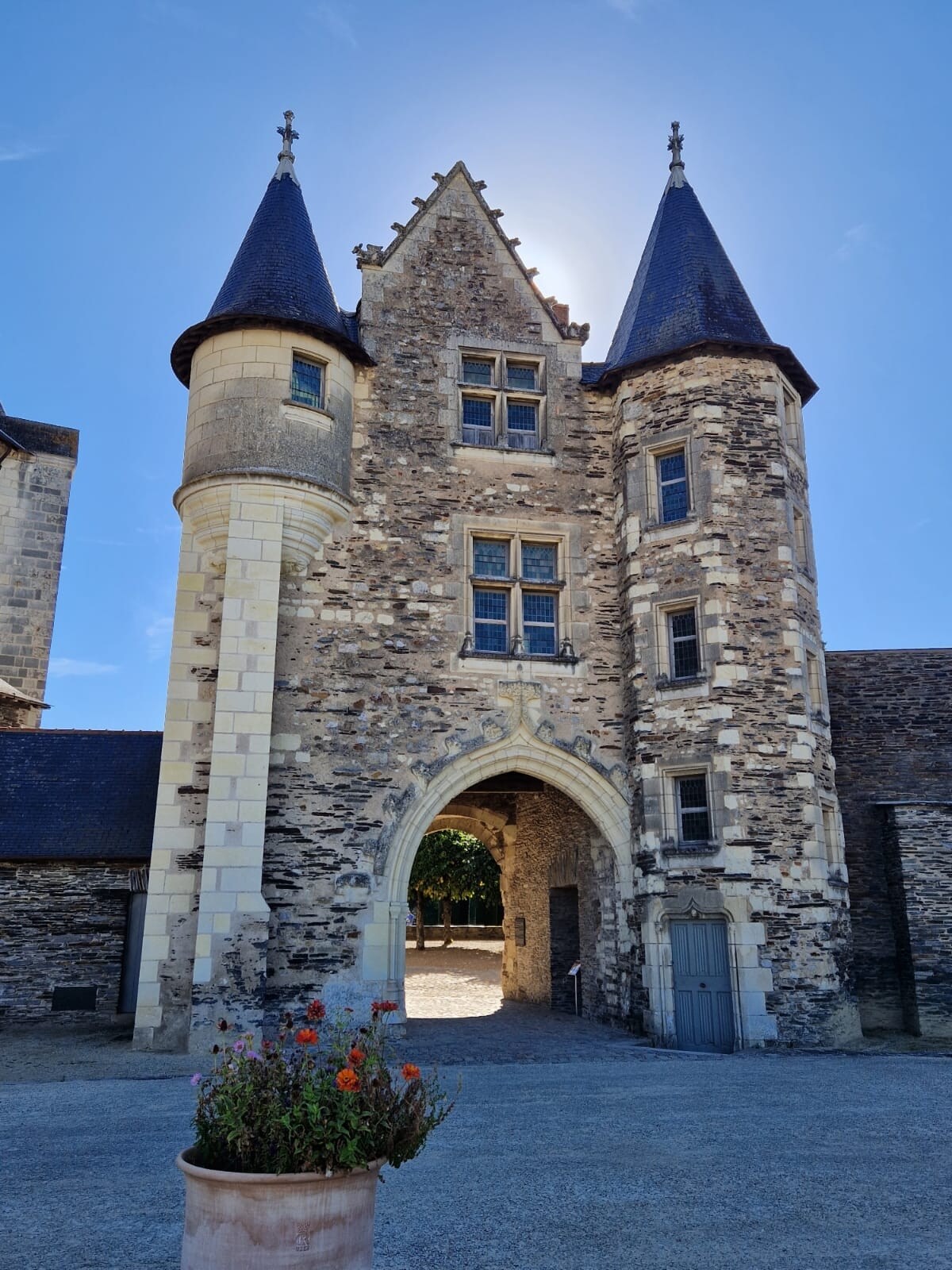 A stone structure with two distinct towers and an arched entrance is featured. The towers have pointed roofs and adorned windows. A paved pathway leads through the archway, while a flower pot filled with vibrant colors is placed prominently at the entrance.