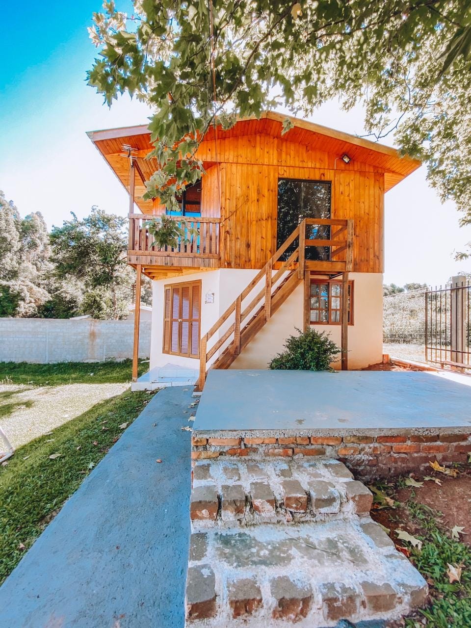 A wooden chalet features a two-story structure with a staircase leading to the upper entrance. The exterior wood finish complements the large windows and the surrounding green lawn. A stone pathway leads to the entrance, framed by trees and open outdoor space.