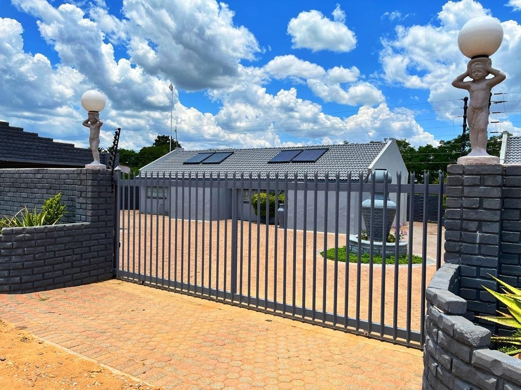 A gated entrance with a stone wall opens to a neatly paved courtyard, featuring a central fountain surrounded by greenery. Solar panels are visible on the roof of the structure, which is flanked by decorative statues on either side of the gate.