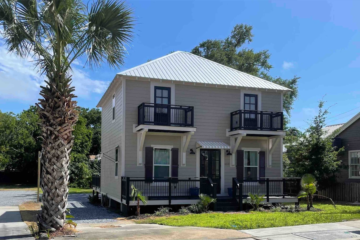 A two-story vacation home with a light exterior and a metal roof is surrounded by greenery. Two balconies are visible on the front, framed by dark shutters. Palm trees are planted nearby, adding a tropical touch to the inviting entrance.