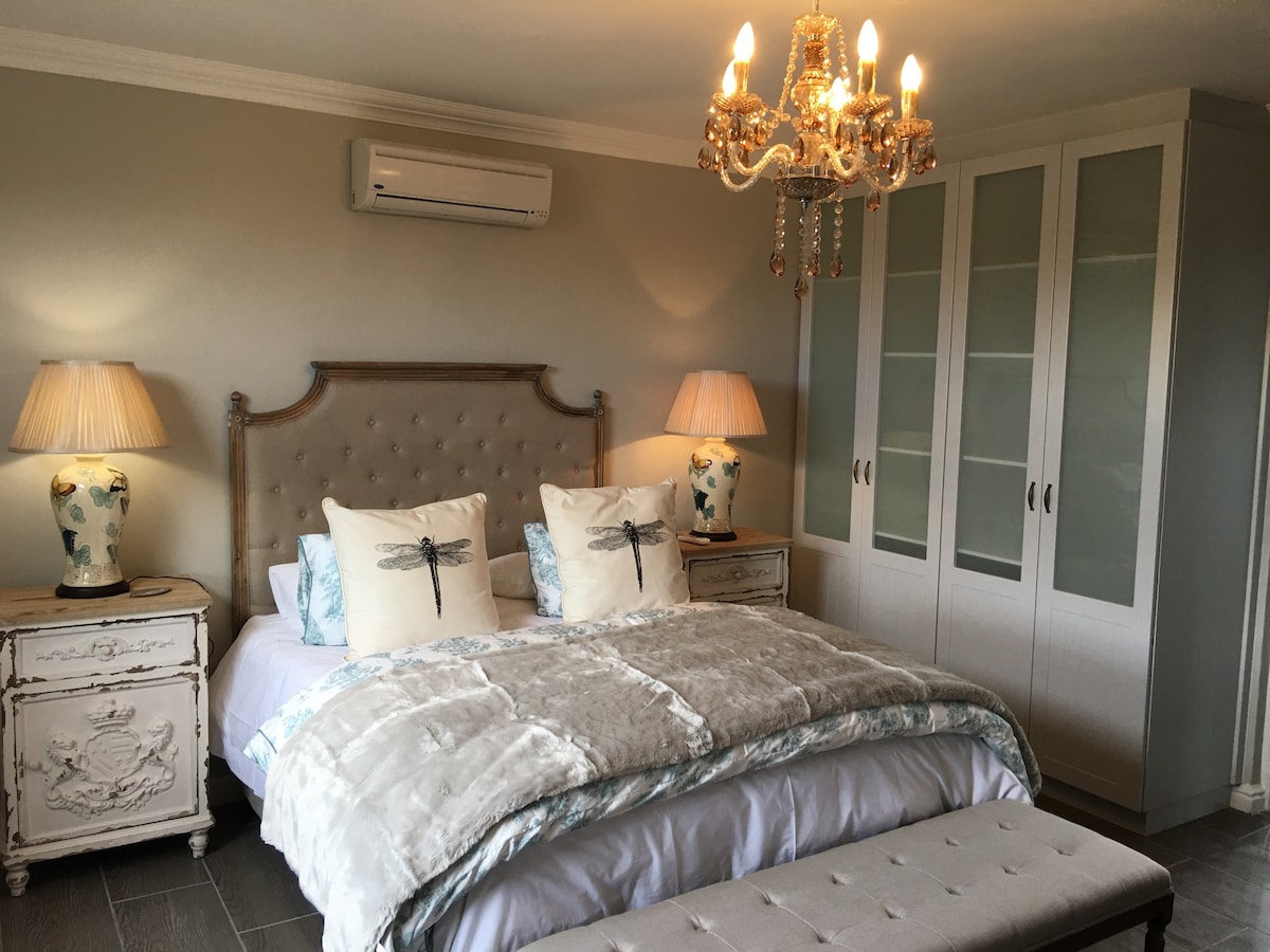 A bright bedroom features a spacious bed adorned with decorative pillows and a plush throw. A stylish chandelier hangs overhead, complemented by matching bedside lamps. An ornate headboard and a light-colored wardrobe are present, adding to the room's elegant design.
