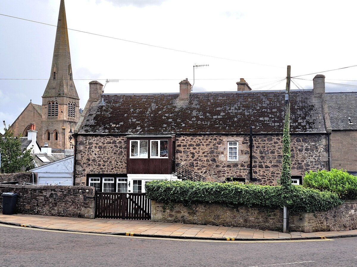 A historic single-level stone building is displayed, featuring a slate roof and a front gate surrounded by a low stone wall. The exterior is accented by climbing greenery. A church steeple is visible in the background, adding to the charming village atmosphere.
