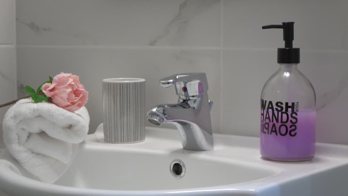 A modern bathroom sink area is presented, featuring a chrome faucet and a decorative soap dispenser with a purple liquid. A neatly rolled white towel and a soft pink rose are placed beside a textured grey vase, creating a clean and inviting space.