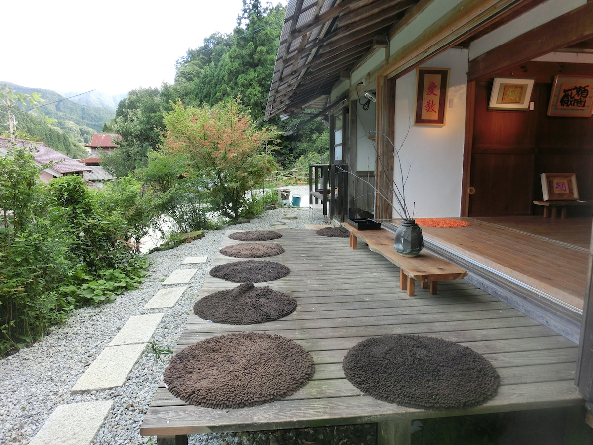 A wooden terrace features circular mats arranged around a simple wooden table. Lush greenery and small stone pathways frame the deck, while a glimpse of a mountain landscape is visible in the distance. Traditional Japanese elements are integrated into the outdoor seating area.