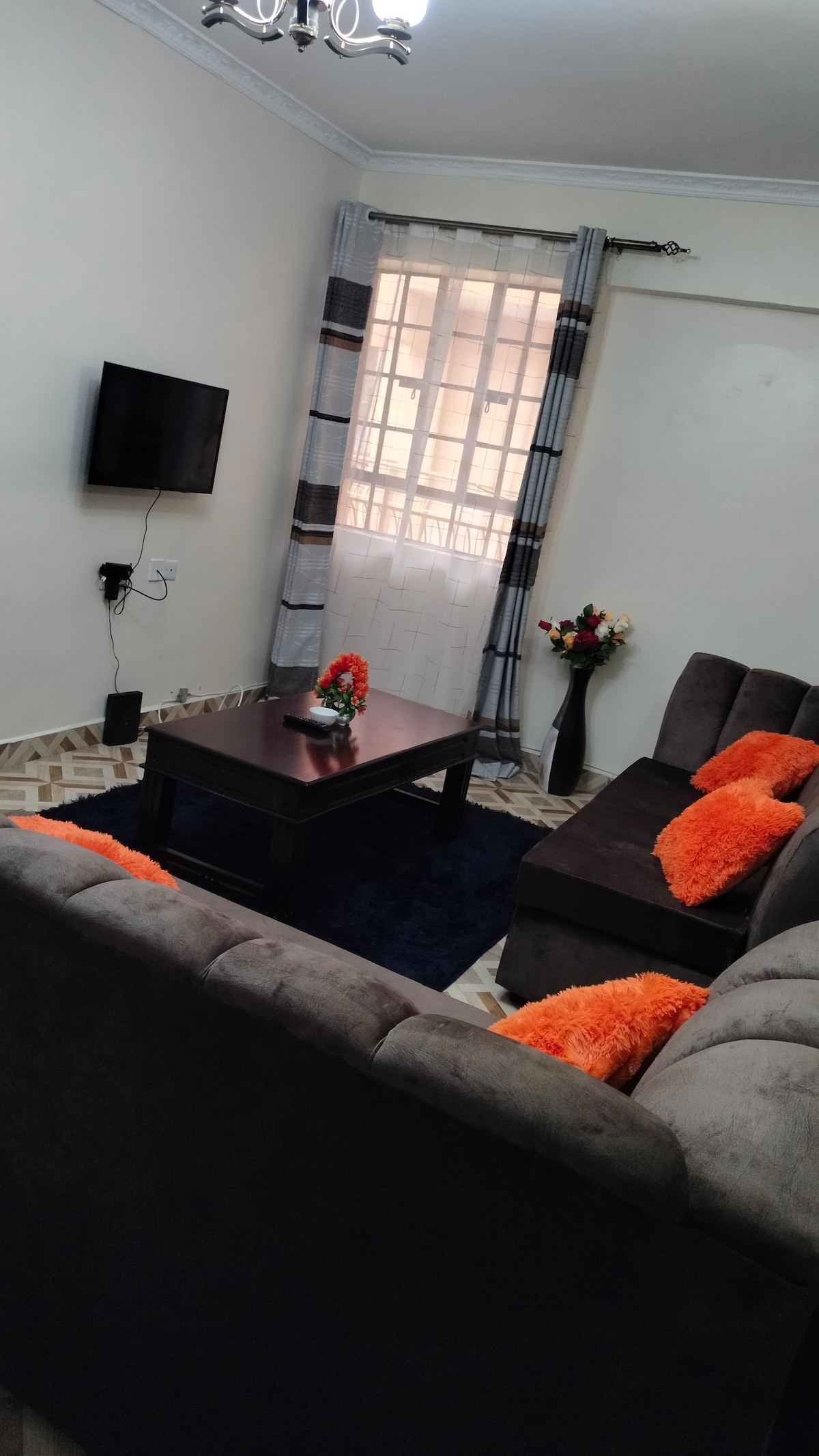 A cozy living area features a dark gray sectional sofa adorned with orange cushions. A low wooden coffee table sits in the center, while a floor vase filled with flowers complements the decor. A wall-mounted television is visible, and sunlight filters through the window with sheer curtains.
