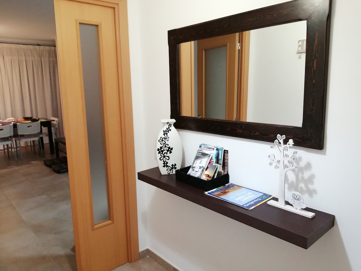 A hallway features a wooden shelf with a decorative vase and a collection of magazines. A large mirror reflects light, creating a sense of space. Light-colored walls complement the tan flooring and lead to a dining area visible in the background.