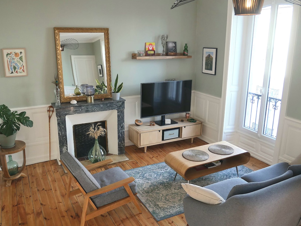 A spacious living area is defined by a vintage fireplace and a large mirror reflecting the soft light. A comfortable couch and a stylish armchair are arranged around a wooden coffee table, with a decorative rug beneath. Decorative plants add a touch of nature.