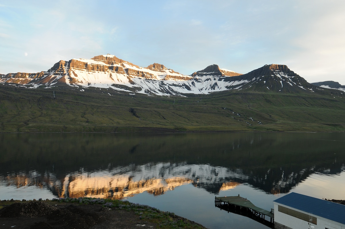 A panoramic view of snow-capped mountains is mirrored in the calm waters of a fjord. The landscape features grassy slopes with patches of dark earth, while a small dock extends into the water, complementing the serene natural setting.