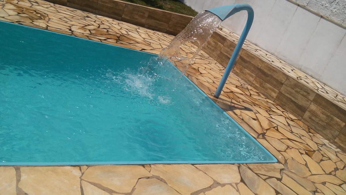 A clear blue swimming pool is depicted with a splash of water flowing from a fountain feature. Surrounding the pool, naturally patterned stone tiles create an inviting area, enhancing the outdoor space. The backdrop includes a smooth white wall that frames the serene environment.