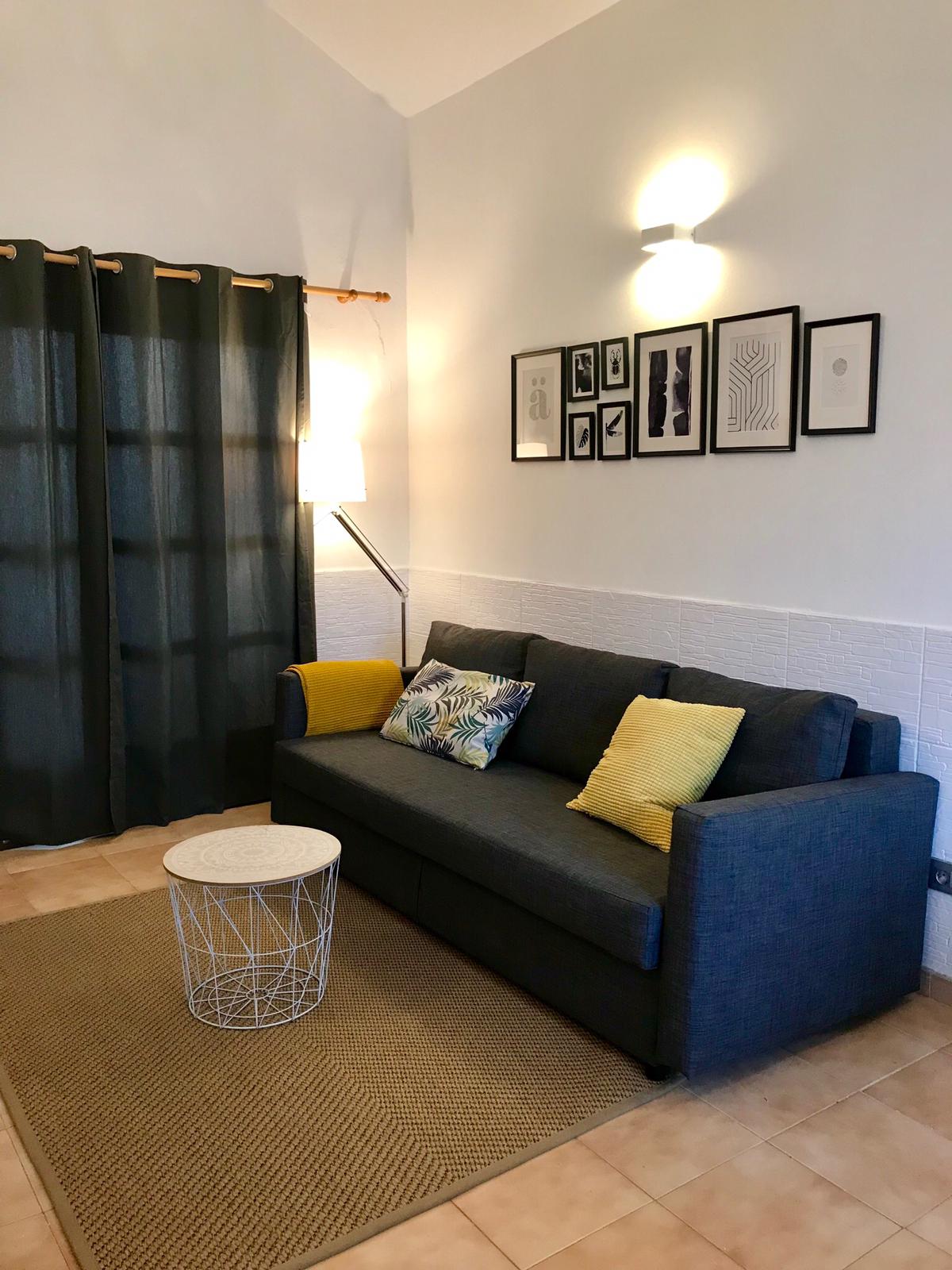 A cozy seating area is presented with a gray sofa adorned with yellow cushions. A small white wire coffee table is positioned in front of the sofa, set atop a textured beige rug. Light is softly diffused through the green curtains hanging over the window.