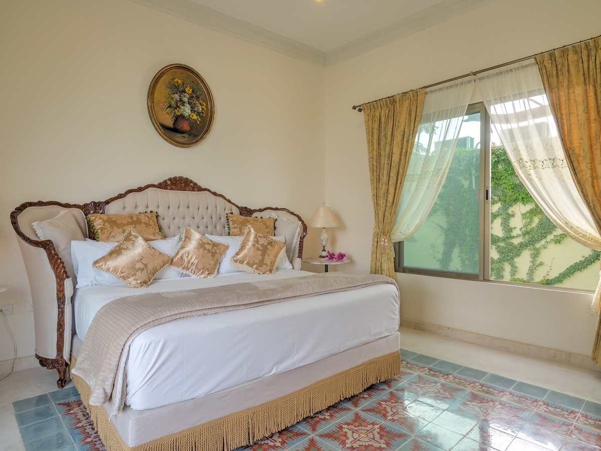A spacious bedroom displays an elegant upholstered headboard and decorative pillows atop a neatly made bed. Natural light filters through sheer curtains, illuminating the room's warm tones and soft furnishings. A tasteful area rug adds color to the tile floor, complementing the overall design.