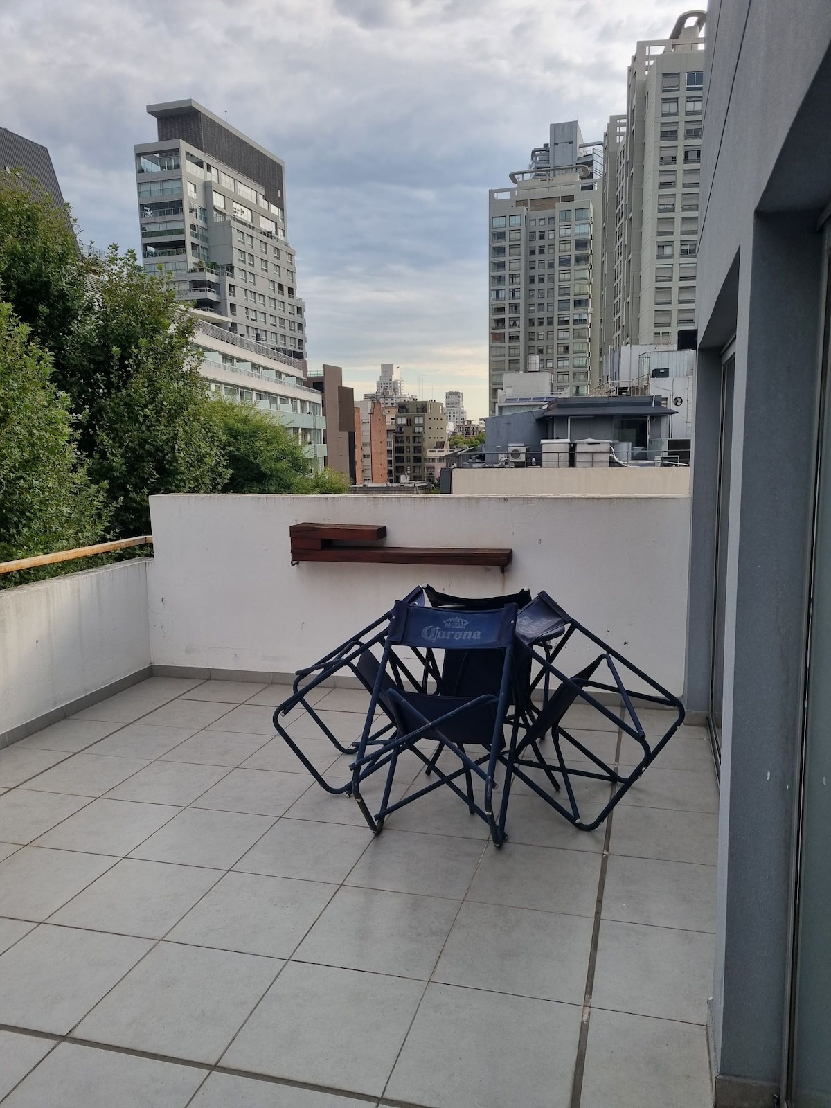 A private balcony is displayed, featuring two folding chairs positioned around a small wooden ledge. The area is surrounded by tall buildings, offering a cityscape backdrop. The tiled floor and greenery provide a sense of openness and connection to the outdoors.